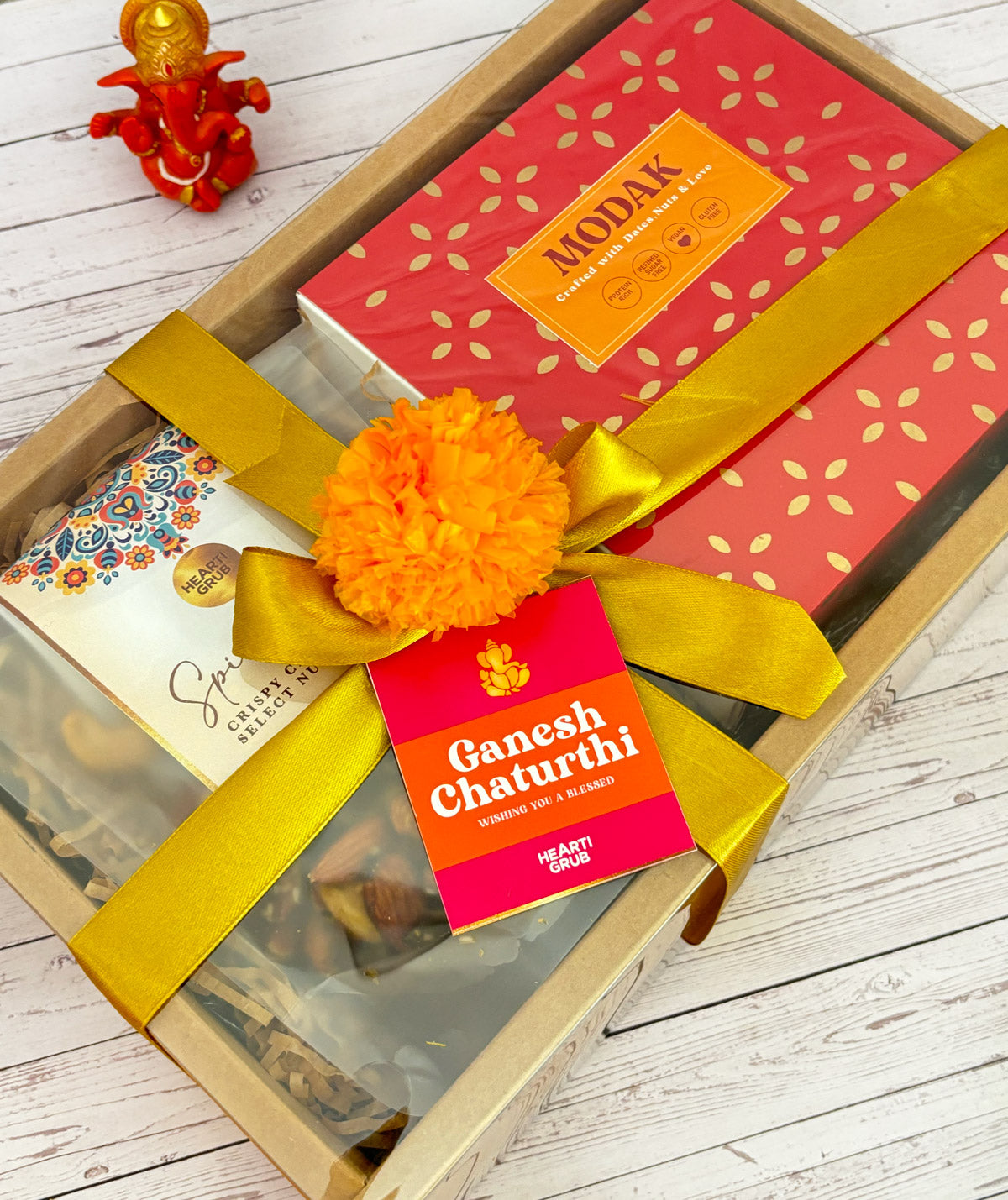 Gift set with Modak, Ganesh Chaturthi items, and a yellow ribbon on a wooden surface. Ganesh Chaturthi. Gifting. Made in Small Batches. Deliciously Decadent. Vegan Gluten Free Refined Sugar Free. Delicious Modak. Nuts. Delivery throughout U.A.E.