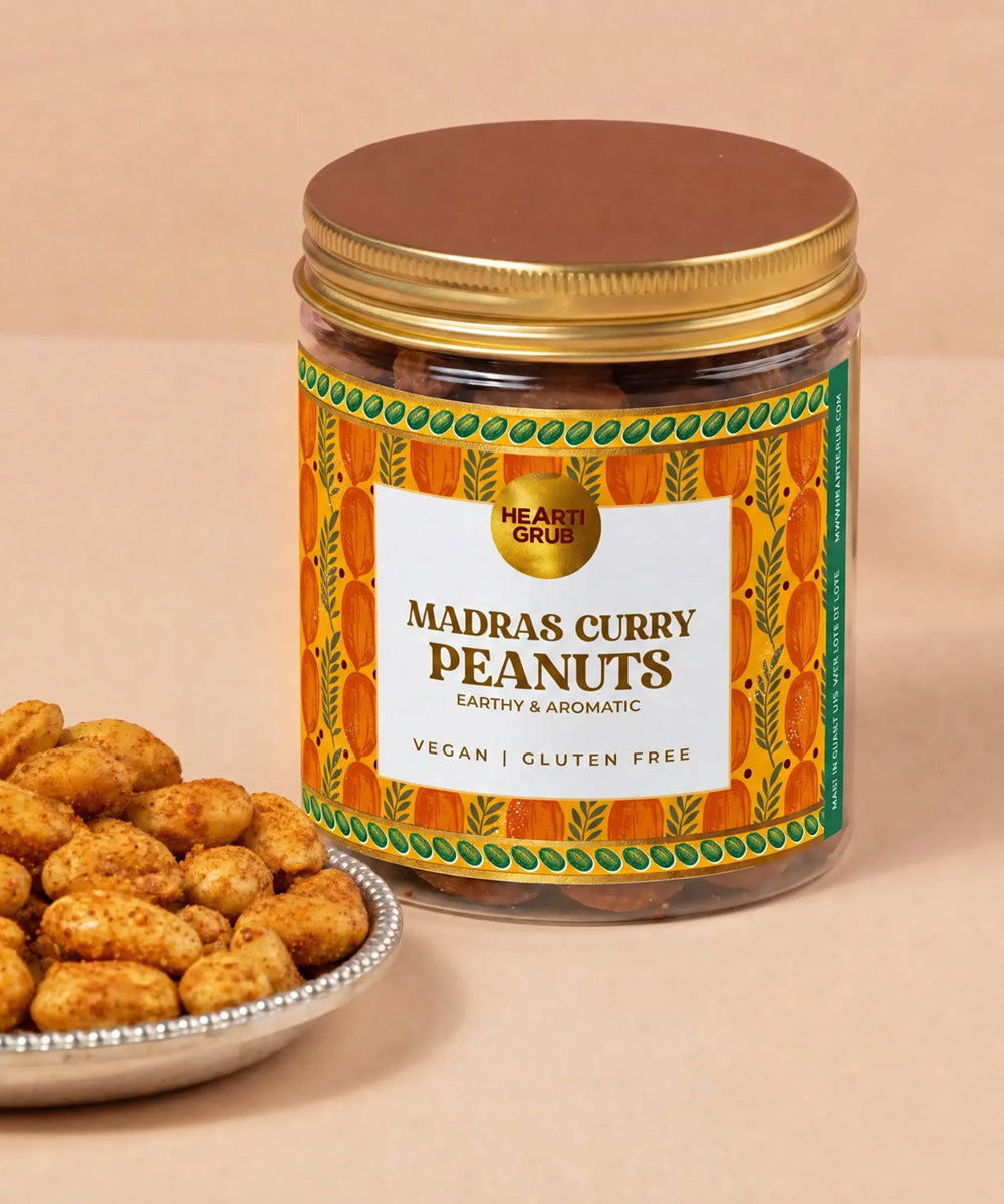 A jar of Hearti Grubs Madras Curry Peanuts with a gold lid and a colorful orange label, shown next to a small plate of roasted peanuts on a beige background. Heartigrub. Diwali Gifting. Dubai, UAE. Abudhabi. Vegan Gifts. Delivery in UAE. Nuts.