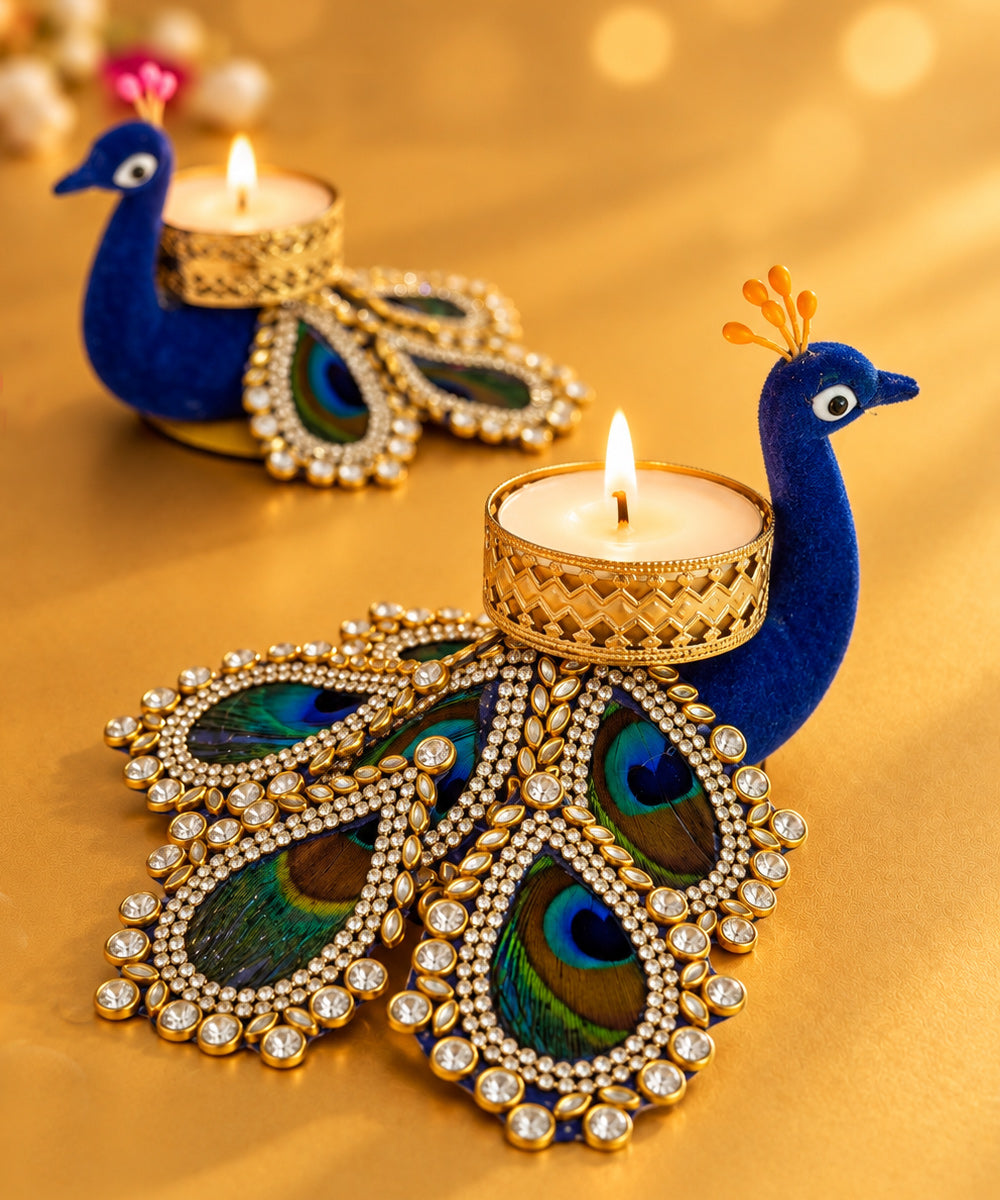 Two ornate peacock-shaped tealight candle holders on a warm gold background. The foreground holder is in sharp focus, showing a blue peacock body, jeweled feather details, and a lit tealight candle in a gold holder. Perfect for: Diwali gifting, luxury hampers, hostess gifts, festive tables & corporate gifting. Heartigrub