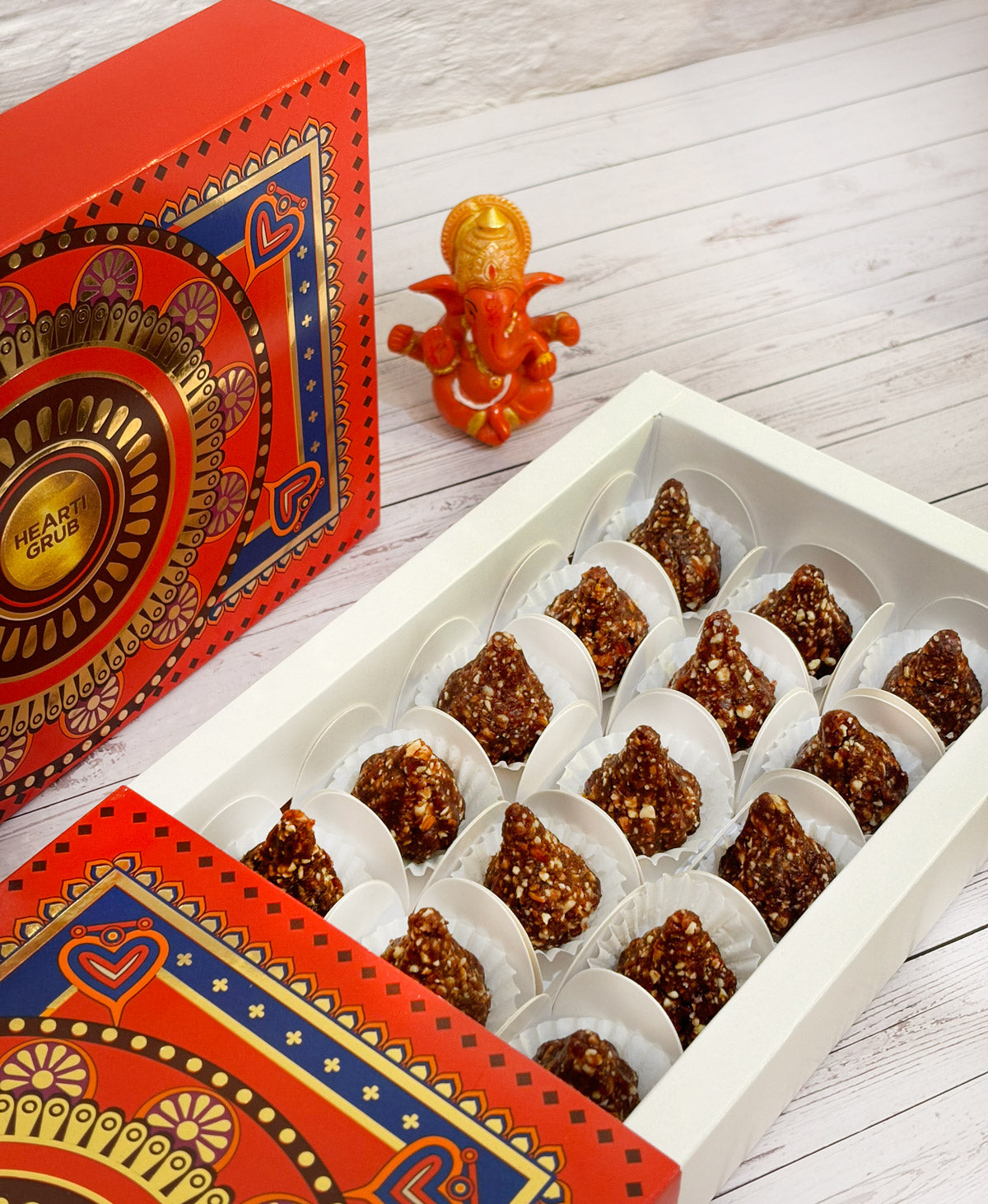 Gift set with Modak, Ganesh Chaturthi items, and a yellow ribbon on a wooden surface. Ganesh Chaturthi. Gifting. Made in Small Batches. Deliciously Decadent. Vegan Gluten Free Refined Sugar Free. Delicious Modak. Nuts. Delivery throughout U.A.E. HeartiGrub