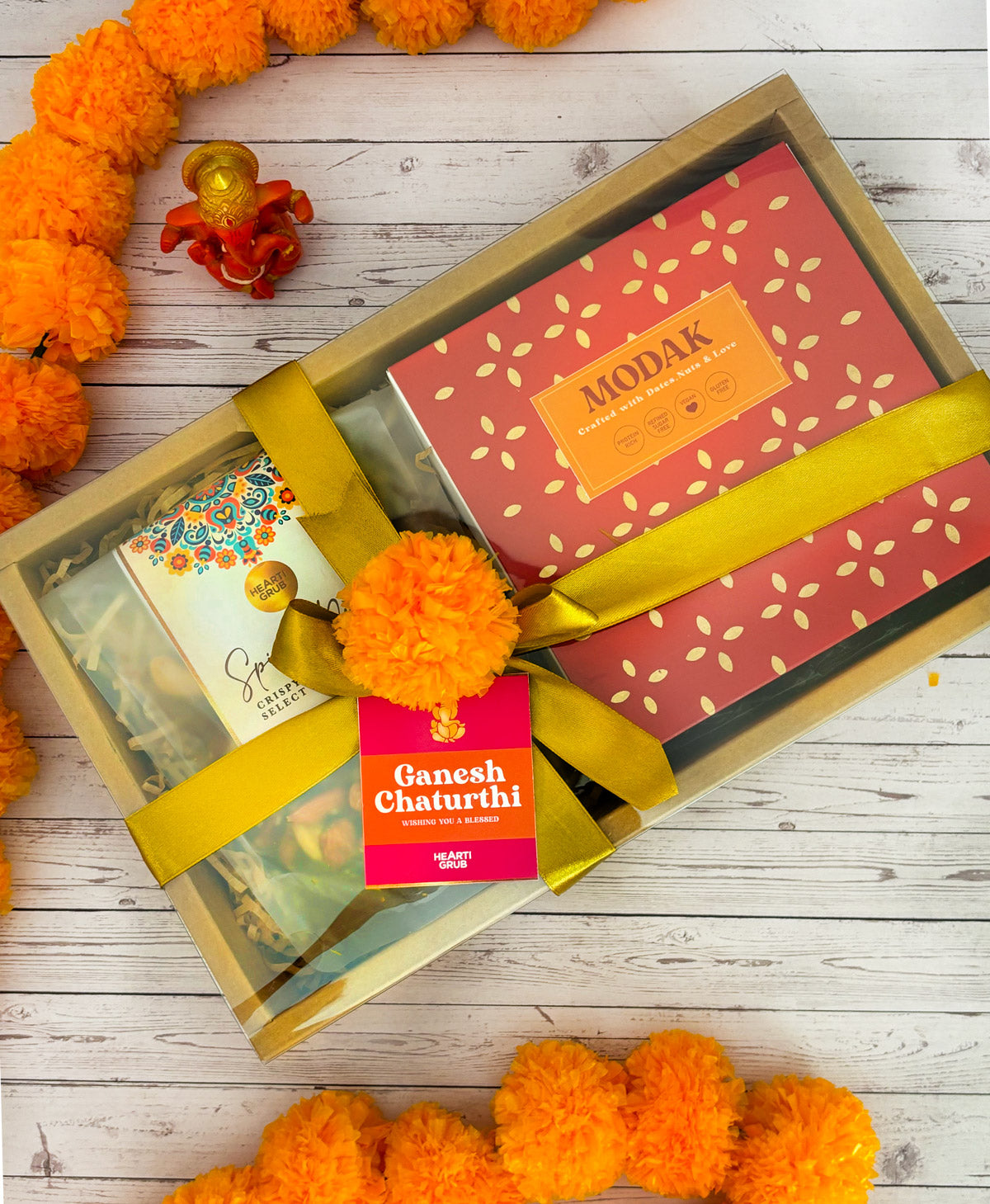 Gift set with Modak, Ganesh Chaturthi items, and a yellow ribbon on a wooden surface. Ganesh Chaturthi. Gifting. Made in Small Batches. Deliciously Decadent. Vegan Gluten Free Refined Sugar Free. Delicious Modak. Nuts. Delivery throughout U.A.E. HeartiGrub