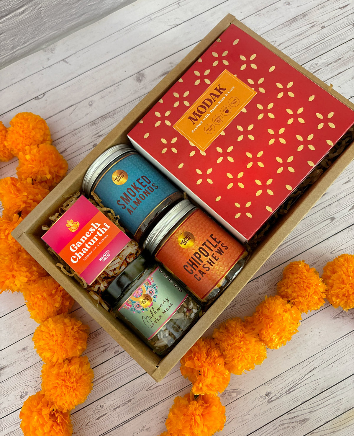 Gift set with Modak, Ganesh Chaturthi items, and a yellow ribbon on a wooden surface. Ganesh Chaturthi. Gifting. Made in Small Batches. Deliciously Decadent. Vegan Gluten Free Refined Sugar Free. Delicious Modak. Nuts. Delivery throughout U.A.E.
