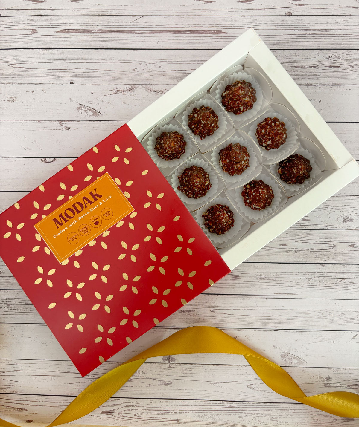 Gift set with Modak, Ganesh Chaturthi items, and a yellow ribbon on a wooden surface. Ganesh Chaturthi. Gifting. Made in Small Batches. Deliciously Decadent. Vegan Gluten Free Refined Sugar Free. Delicious Modak. Nuts. Delivery throughout U.A.E. HeartiGrub