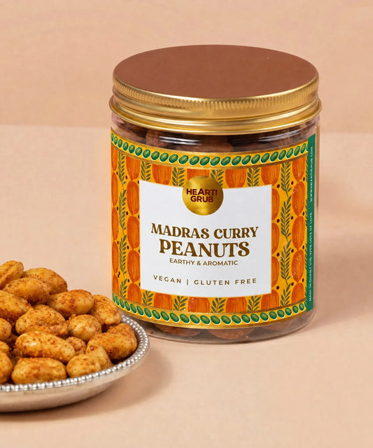 A jar of Hearti Grubs Madras Curry Peanuts with a gold lid and a colorful orange label, shown next to a small plate of roasted peanuts on a beige background. Heartigrub. Diwali Gifting. Dubai, UAE. Abudhabi. Vegan Gifts. Delivery in UAE. Nuts.