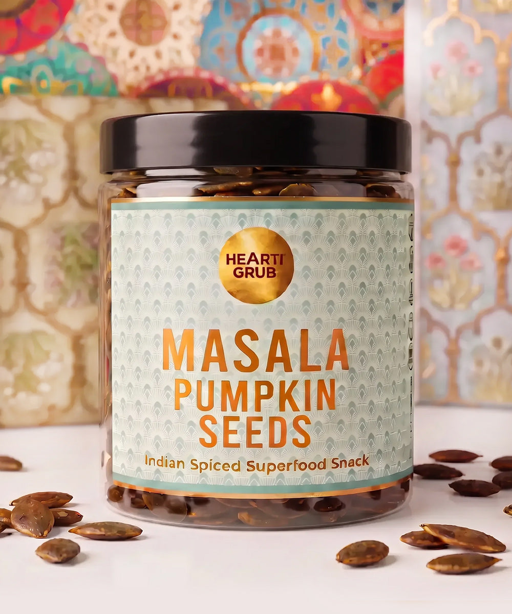 Diwali Gifts. Dubai. UAE. heartiGrub. nuts and Seeds. Snacks. Clean Snacking. A clear plastic jar of Hearti Grub Masala Pumpkin Seeds with a black lid and a light green patterned label, set on a white surface with scattered pumpkin seeds and a colorful patterned background.