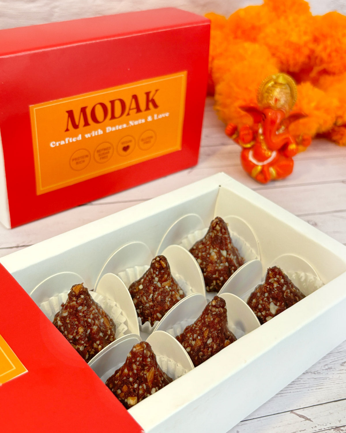 Gift set with Modak, Ganesh Chaturthi items, and a yellow ribbon on a wooden surface. Ganesh Chaturthi. Gifting. Made in Small Batches. Deliciously Decadent. Vegan Gluten Free Refined Sugar Free. Delicious Modak. Nuts. Delivery throughout U.A.E. HeartiGrub
