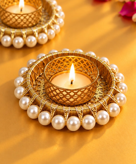 Moti diya Dubai · pearl diya Dubai · Diwali diya UAE · luxury Diwali gifts Dubai · Diwali gift add-ons · festive décor Dubai · premium Diwali hampers UAE · corporate Diwali gifts Dubai · decorative diya UAE · unique Diwali gifts Dubai. Heartigrub. A close-up of a gold-plated decorative tea light holder adorned pearls, holding a lit candle, set against a warm golden background with another similar holder blurred in the background. 